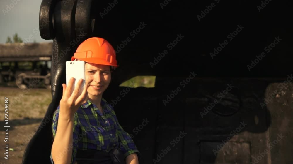 A young woman worker in an orange helmet sits in a large bucket of a mine excavator and take a selfie using a white smartphone camera.