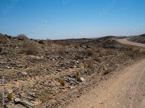 Adventure journey scene on dirt road trip through Namib desert hot dried landscape to rock mountain horizon with local plant, splitting stone and clear blue sky