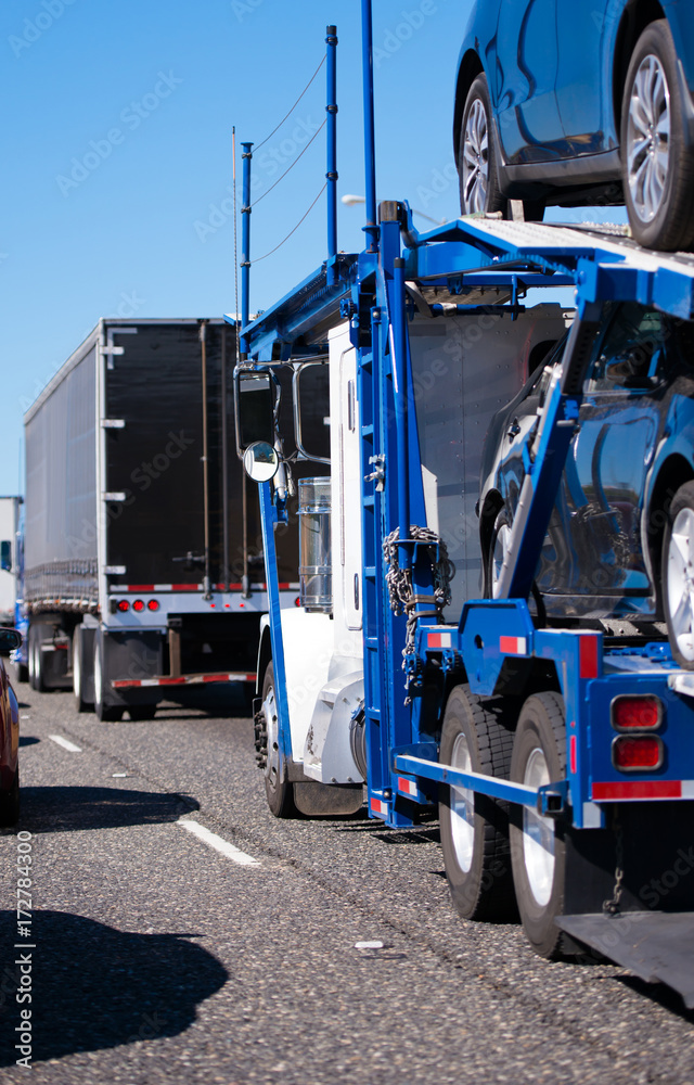 Big rig semi truck with car hauler trailer transporting cars on busy highway with heavy traffic in convoy with another semi trucks Big rig semi truck with car hauler trailer transporting cars on busy highway with heavy traffic in convoy with another semi trucks
