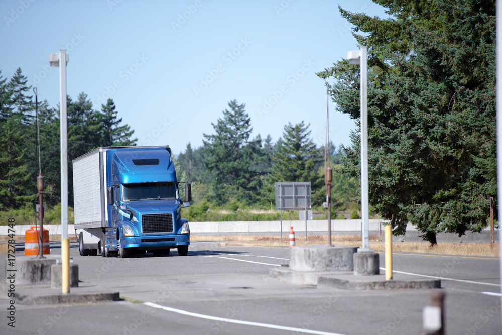 Blue modern semi truck with trailer running to rest area entrance with green trees