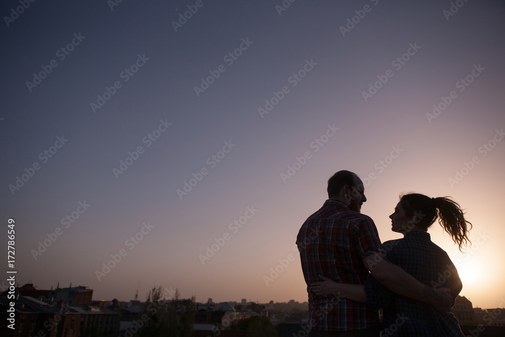 Love couple in beautiful sunset. Cuddling people on roof in focus on foreground. Tender look, creative date, hipster family, intimate relationship, soul mate concept. Urban background with free space