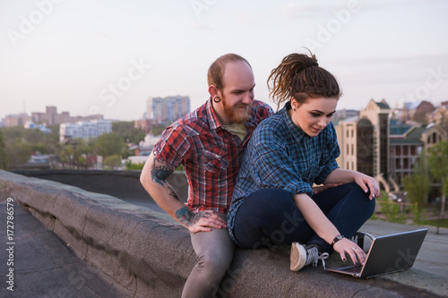 Modern hipster youth couple on roof. Social media activity outdoors. Creative couple leisure. Urban background, smiling young people with laptop