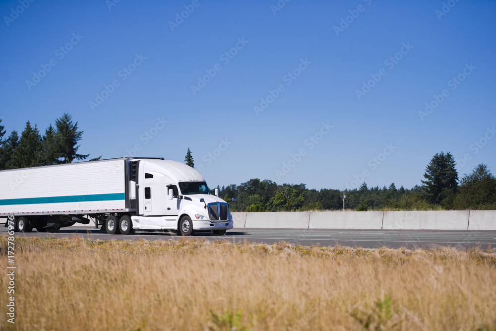 Modern white big rig semi truck with reefer trailer running on highway with yellow grass on shoulder