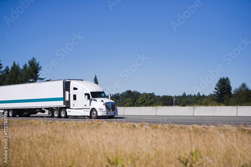Modern white big rig semi truck with reefer trailer running on highway with yellow grass on shoulder