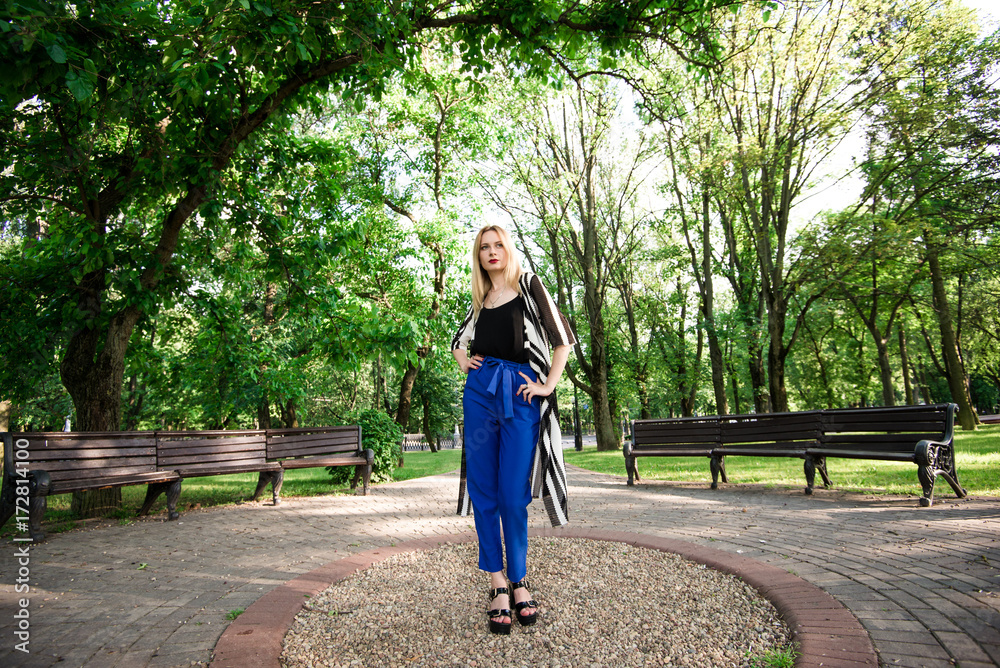 Девушка со светлыми волосами стоит в парке в синих брюках и черной майке. Стильная девушка 