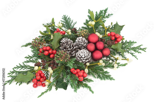 Christmas centrepiece decoration with red baubles, holly, ivy, mistletoe, cedar and juniper leaf sprigs and pine cones on white background.
