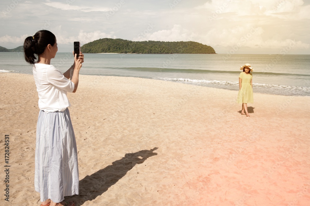 Young asian woman standing on the beautiful nice blue beach with white sand and taking photo with smartphone. Young asian woman standing on the beautiful nice blue beach with white sand and taking photo with smartphone.