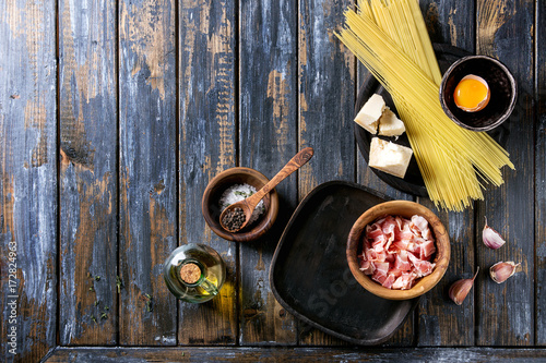 Ingredients for traditional italian pasta alla carbonara. Uncooked spaghetti, pancetta bacon, parmesan cheese, egg yolk, salt, pepper in olive wood bowls over old plank background. Top view with space photo