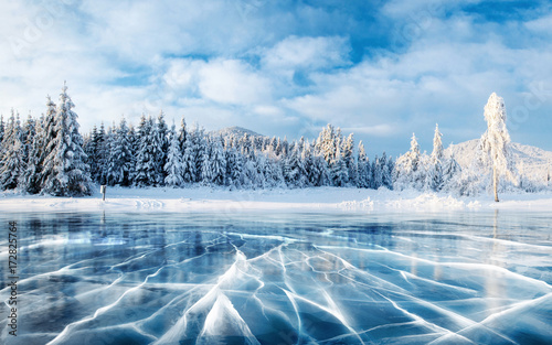 Niebieski lód i pęknięcia na powierzchni lodu. Zamarznięty jezioro pod niebieskim niebem w zimie. Wzgórza sosen. Zimowy. Karpackie, Ukraina, Europa.