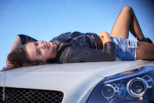 sexy girl in a leather jacket, denim shorts and black pantyhose in the net lies on the hood of the car