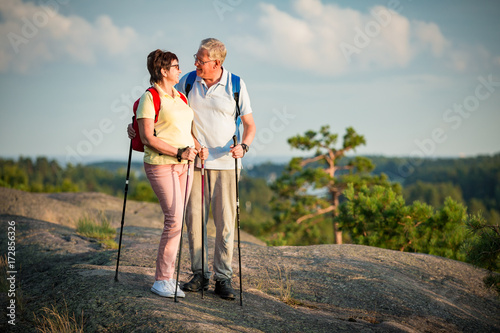 Active senior couple hiking on the top of rock, exploring. Mature man and woman Happily smiling. Nordic walking, trekking. Scenic view of gulf and sea. Healthy lifestyle. Finland.