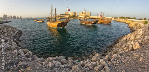 Traditional wooden boats dhow in Qatar in daylight with stones in foreground and Doha skyline in background.	 photo