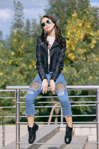 fashionable young woman in rock style clothes, black leather jacket, blue jeans, tights in a grid under battered jeans