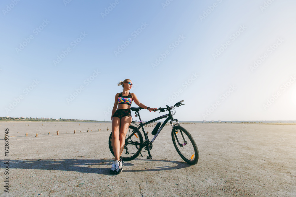 Fototapeta premium A strong blonde woman in a multicolored suit and sunglasses stands near a bicycle in a desert area and looks at the sun. Fitness concept. Blue sky background
