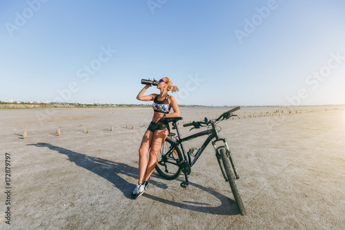 Fototapeta Naklejka Na Ścianę i Meble -  A strong blonde woman in a multicolored suit and sunglasses stands near a bicycle, drinks water from a bottle in a desert area. Fitness concept. Blue sky background