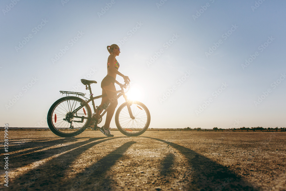 Fototapeta premium A silhouette of a strong woman in a colorful suit that sits on a bicycle in a desert area. Fitness concept. Blue sky background