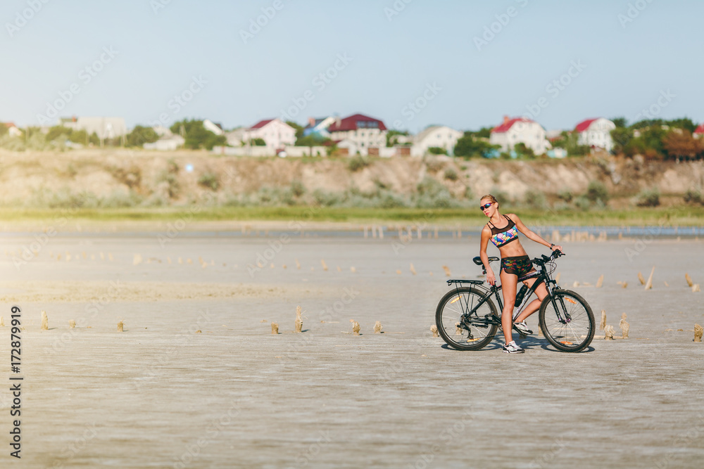 Obraz premium A strong blonde woman in a multicolored suit and sunglasses sits on a bicycle in a desert area. Fitness concept. Blue sky background