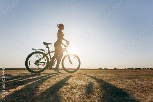 A silhouette of a strong woman in a colorful suit that sits on a bicycle in a desert area. Fitness concept. Blue sky background