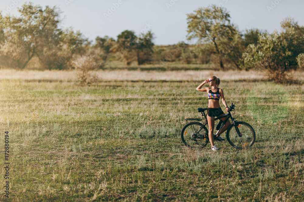 A strong blonde woman in a colorful suit sits on a bicycle in a desert area with trees and green grass and looks at the sun. Fitness concept.