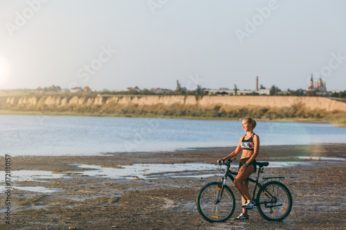 A strong blonde woman in a colorful suit sits on a bicycle in a desert area near the water and looks at the sun. Fitness concept. Blue sky background