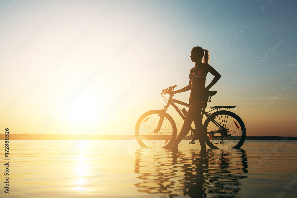 Fototapeta premium A strong blonde woman in a colorful suit stands near the bicycle in the water at sunset on a warm summer day. Fitness concept. Sky background