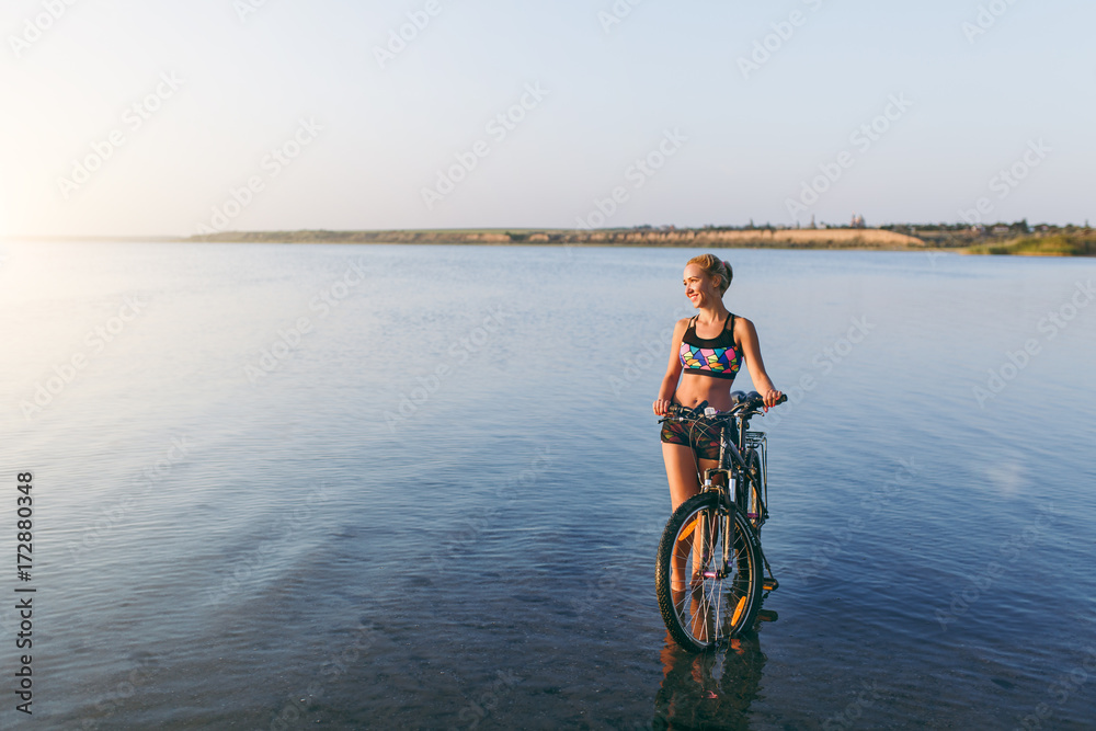 A strong blonde woman in a colorful suit stands near the bicycle in the water at sunset on a warm summer day. Fitness concept. Sky background