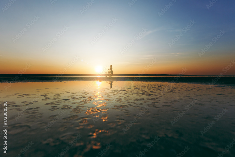 Silhouette of a sporty girl in a suit standing near a bicycle in the water at sunset on a warm summer day. Fitness concept. Sky background