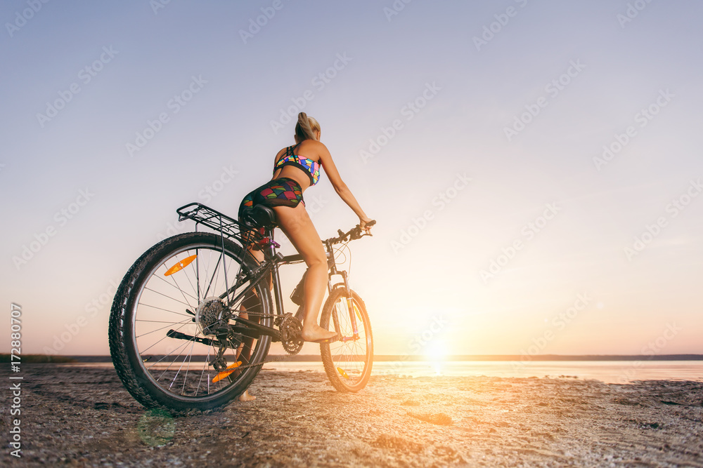 Fototapeta premium A strong blonde woman in a multicolored suit sits on a bicycle in a desert area near the water and looks at the sun. Fitness concept. Back view