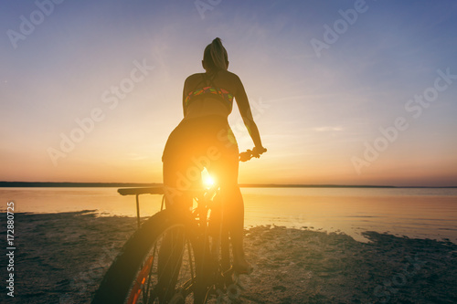 A strong blonde woman in a multicolored suit sits on a bicycle in a desert area near the water and looks at the sun. Fitness concept. Back view © ViDi Studio