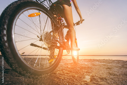 A woman in a multicolored suit sits on a bicycle in a desert area near the water. Fitness concept. Rear view and bottom view. Close-up