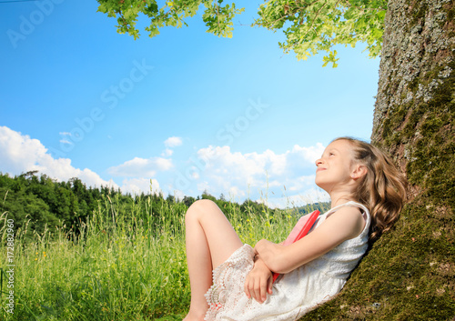 Fotografie young girl sit next to a tree and smile into the summer sun