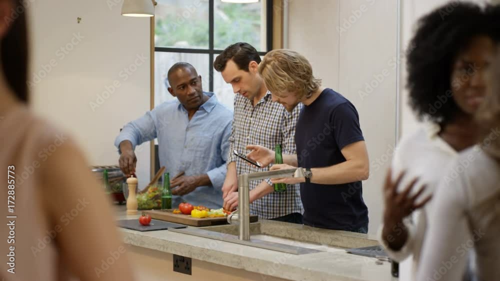 Happy mixed ethnicity group of friends socializing at home. Preparing a meal, chatting and looking at smartphone.