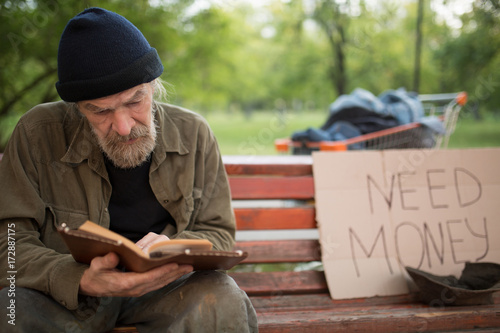 Old beardy man with no home reading a book. Homeless male reading, begging for money on card board.