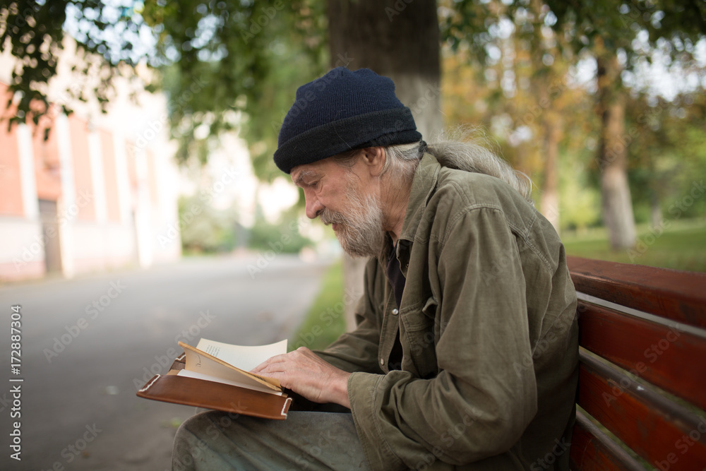 Side view of old homeless man with long grey hair reading a book. Tramp sitting on the bench in the street,