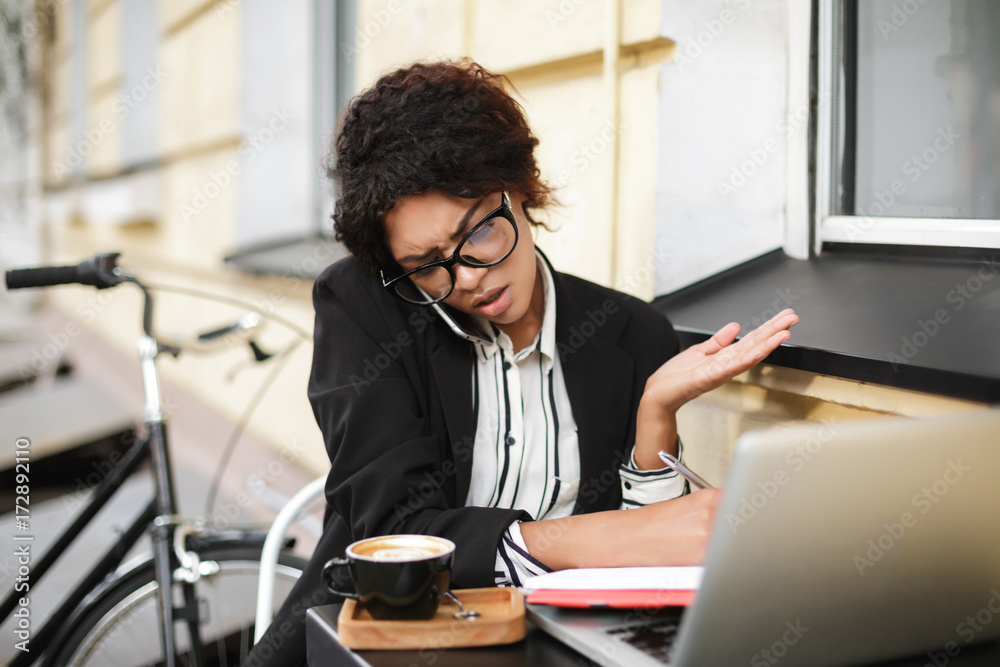 Portrait of African American girl in glasses sitting at the table and emotionally talking on her cellphone. Lady with dark curly hair amazedly looking aside with laptop on table and bicycle nearby