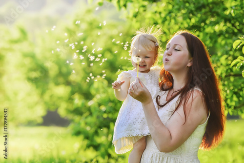 woman and child blowing on a dandelion against a background of green trees with shallow depth of field