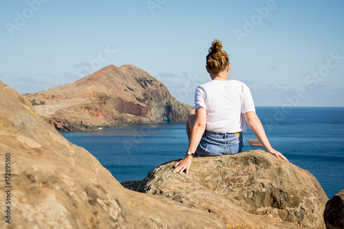 Junge Frau mit einer Dutt-Frisur sitzt auf einem Felsen und blickt in Richtung Meer © Remo