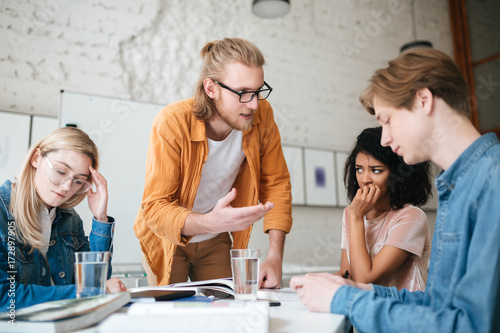 Portrait of teacher with blond hair and beard leaning on table and emotionally explaining something to students. Group of upset people sitting at the table and working together with books