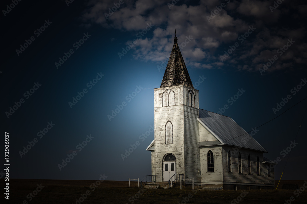 horizontal image of a bright light shining on an old white country church in the late evening and night time.
