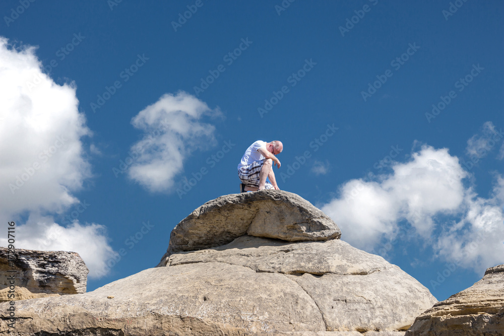 horizontal image of a middle aged caucasian man sitting on a tall boulder in the summer time taking time for solitude under a bright blue sky with white clouds floating by in the summer time horizontal image of a middle aged caucasian man sitting on a tall boulder in the summer time taking time for solitude under a bright blue sky with white clouds floating by in the summer time