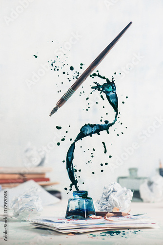 Spilled ink flying above inkwell in a spiraling splash with tiny drops and flying pen on a light background. Still life with writer workplace. Creative writing concept. photo