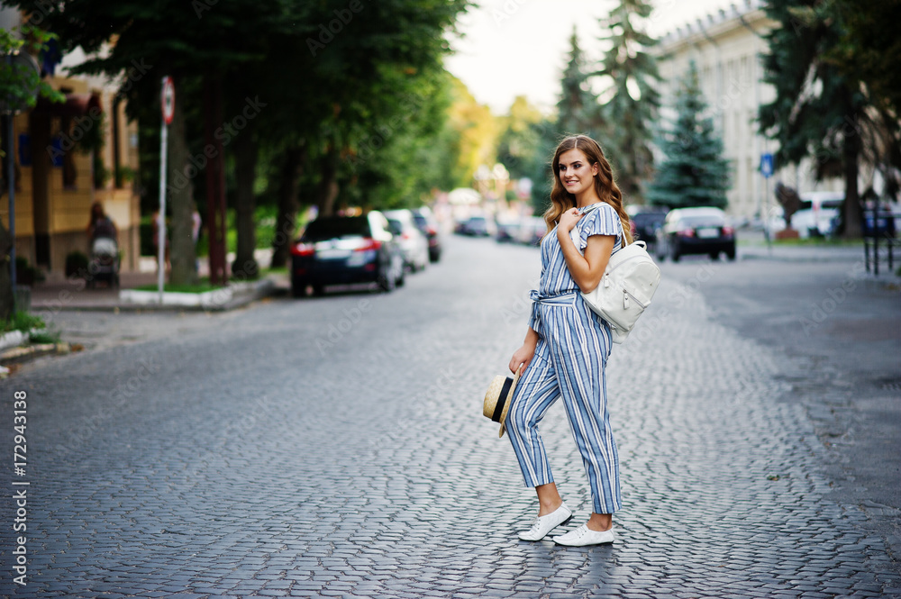 Portrait of a beautiful model in striped overall posing with hat and a backpack on a street with trees in a town.