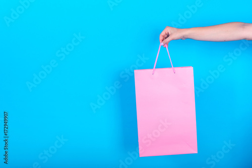Female hand holding pink shopping bags isolated on blue (cyan) background, free space, close up. Sale, advertisement and black friday concept. Closeup of woman hand carries shopping bag