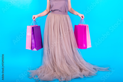 Beautiful young slim woman in long dress holding shopping bags over blue (cyan) background, copy space. Beauty, fashion, seasonal sale concept. Black friday, free space