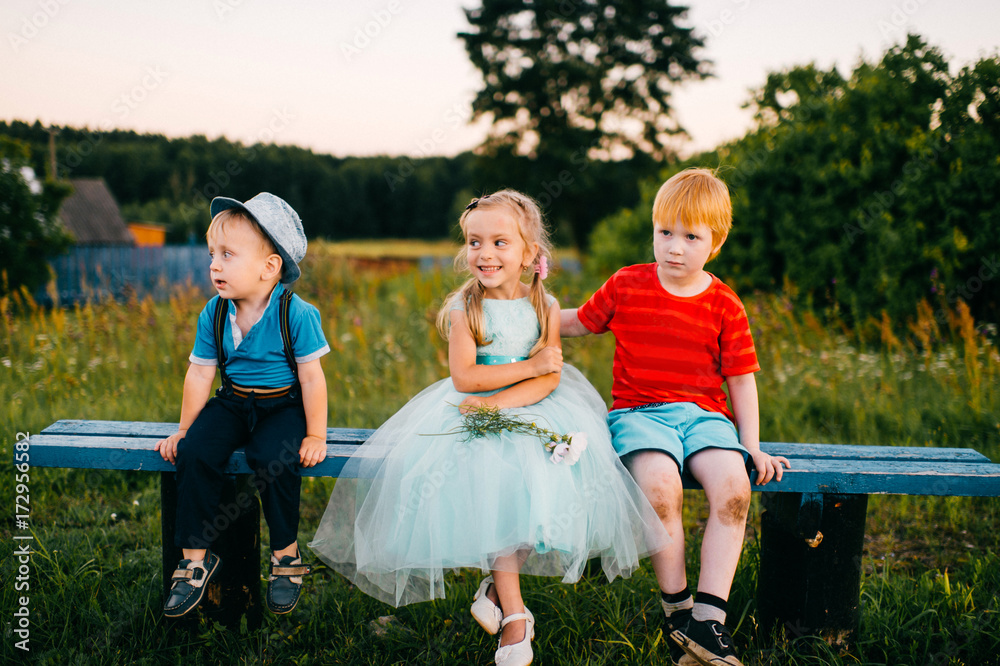 Fototapeta premium Group of little emotional kids sitting on bench outdoor in countryside. Girl in dress between two guys. Difficult relations. Youth jealousy. Funny loving triangle. Joy, sorrow, hurt and offence.