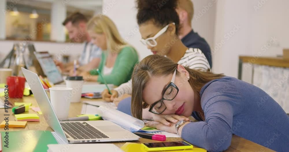 Woman in glasses lying on table in office and sleeping on background of young colleagues working with documents.