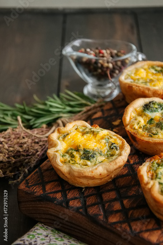 Savory mini quiches (tarts) on a wooden board. Flaky dough pies. Fresh rosemary and dry thyme on a wooden background.