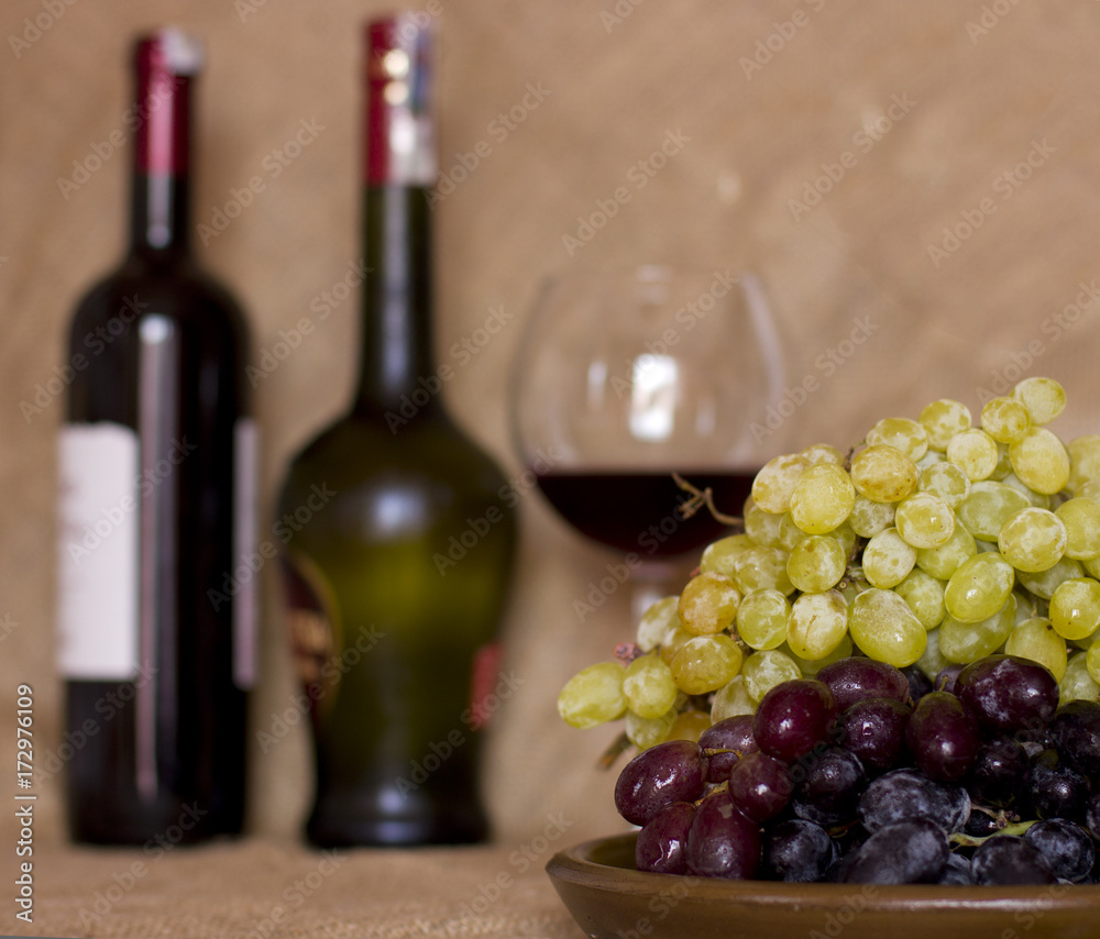 Blue and green grapes on a clay brown dish. Bottle with red and white wine on background