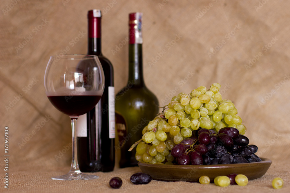 Blue and green grapes on a clay brown dish. Bottle with red and white wine on background