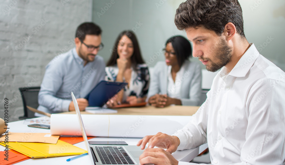 Young handsome businessman working on laptop with coworkers discussing a new project in the background. Young handsome businessman working on laptop with coworkers discussing a new project in the background.